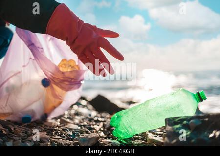 Tag Der Erde. Eine Person mit schützenden Gummihandschuhen greift nach einer schmutzigen grünen Plastikflasche, die am Meeresufer liegt. Nahaufnahme der Hand. Speicherplatz kopieren. Konzept Stockfoto