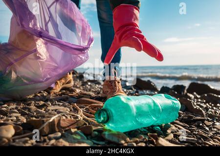 Ein Freiwilliger in Gummihandschuhen greift nach einer schmutzigen Plastikflasche, die am Meeresufer liegt. Nahaufnahme der Hand. Das Konzept der Umweltreinigung Stockfoto