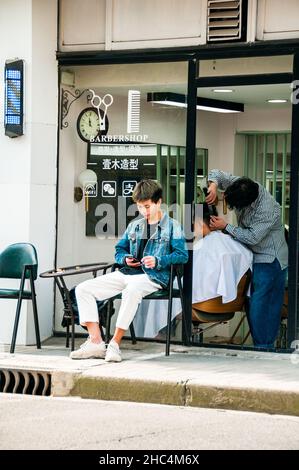 Ein Barbershop in den Straßen um 1933 im Bezirk Hongkou mit einem jungen Barbier draußen, der sein Telefon benutzte. Shanghai, China. Stockfoto