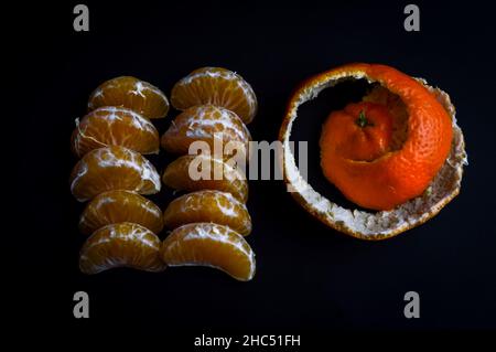 Organische Mandarinenkeile Reihen sich mit der Schale in einer Spirale neben ihnen auf einem schwarzen Papierhintergrund zusammen Stockfoto