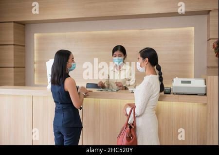 Asiatische Patientin mit Gesichtsmaske, Schwester steht mit Rezeptionistin am Schalter in der Klinik in Kontakt Stockfoto