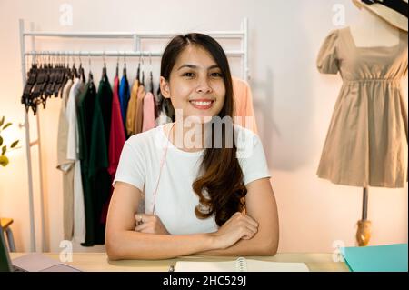 Glückliche asiatische Frau Unternehmerin von Online-Bekleidungsgeschäft sitzen und gekreuzte Arme auf dem Schreibtisch im Home Office Stockfoto