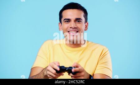 KIEW, UKRAINE - 22. DEZEMBER 2021: Fröhlicher junger Mann mit Joystick und Videospiel isoliert auf Blau Stockfoto