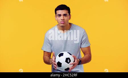 Besorgter junger Mann im T-Shirt, der Fußball isoliert auf Gelb hält Stockfoto