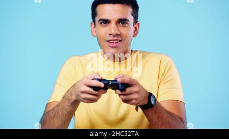 KIEW, UKRAINE - 22. DEZEMBER 2021: Glücklicher junger Mann mit Joystick und Videospiel isoliert auf Blau Stockfoto