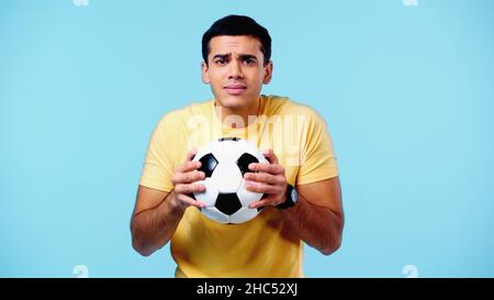 Ängstlicher junger Mann in gelbem T-Shirt, der Fußball isoliert auf Blau hält Stockfoto