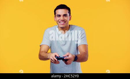 KIEW, UKRAINE - 22. DEZEMBER 2021: Fröhlicher junger Mann mit Joystick und Videospiel isoliert auf Gelb Stockfoto