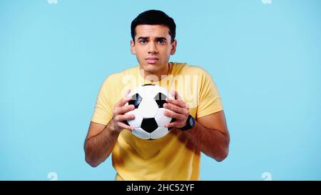 Gestresster junger Mann in gelbem T-Shirt, der Fußball isoliert auf Blau hält Stockfoto