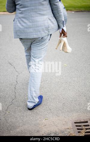 Vertikale Aufnahme eines Bräutigams, der auf der Autobahn läuft und in der Hand weiße Schuhe der Braut hält Stockfoto