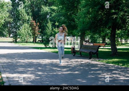 Laufen schnell lächelnd junge sportliche Frau wärmen sich auf, Stretching in grau Sportkleidung im Park an sonnigen Sommertag Stockfoto