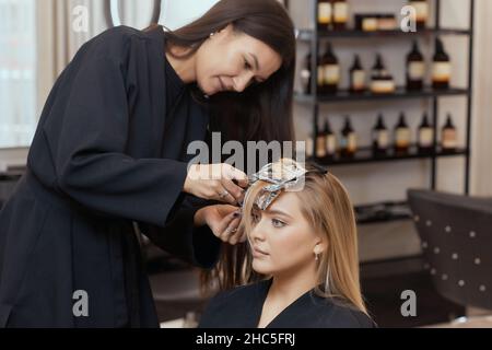 Friseur macht Bouffant mit Kamm auf dünnen Strähnen. Shatush Technik für Haaraufhellung Stockfoto