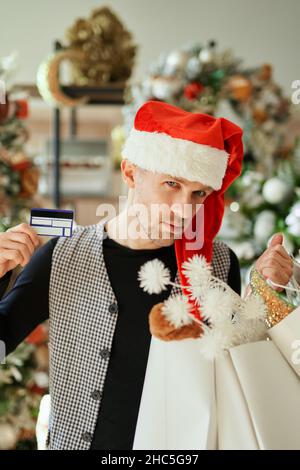 Kaukasischer Mann mit Einkaufstaschen und Kreditkarte, trägt Weihnachtsmütze auf dem Einkaufszentrum oder Weihnachtsdekoration Hintergrund. Spezielles Weihnachtsangebot für Kunden. Weihnachtsverkauf und Shopping-Konzept Stockfoto