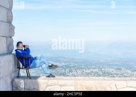 Lovcen, Montenegro-September 14 2019: Auf dem Gipfel des Mount Lovcen, an einem warmen sonnigen Nachmittag, sitzt eine Person auf einer Wand in seinem Stuhl und blickt hinunter auf die Stockfoto