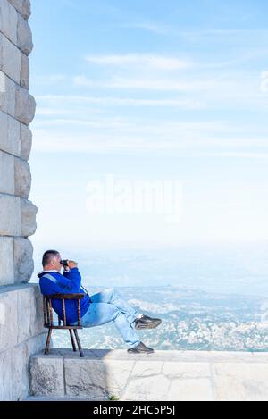 Lovcen, Montenegro-September 14 2019: Auf dem Gipfel des Mount Lovcen, an einem warmen sonnigen Nachmittag, sitzt eine Person auf einer Wand in seinem Stuhl und blickt hinunter auf die Stockfoto