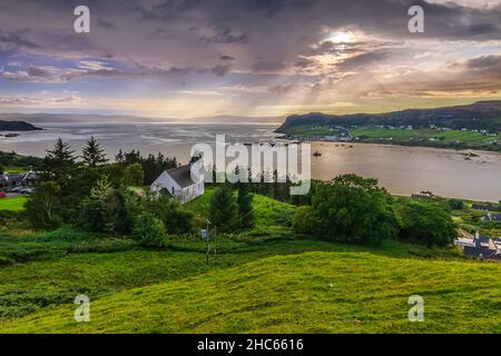 Dramatische Wolken am Himmel bei Sonnenuntergang in Schottland auf der Isle of Skye. Einsame Kirche auf einem Hügel mit grüner Wiese und Bäumen. Sonnenstrahlen am Horizont. Sm Stockfoto