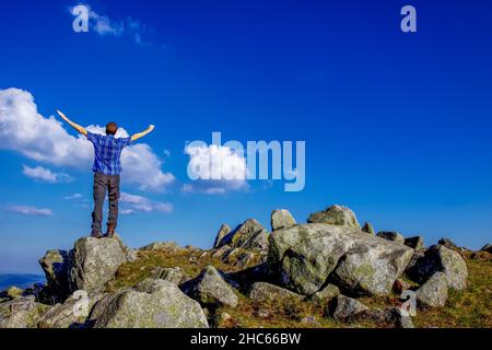 Ein einzelner Mann steht mit ausgestreckten Armen auf einem Felsen vor einem blauen Himmel Stockfoto