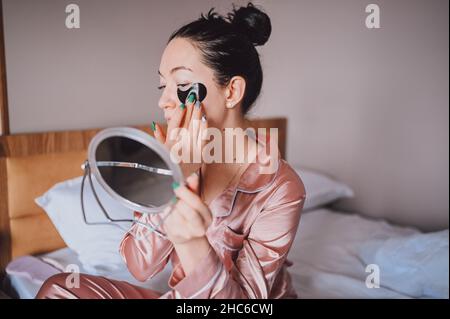 Kosmetik, Hautpflege, Gesichtsbehandlung, Spa, natürliches Schönheitskonzept. Lächelnde, fröhliche junge Frau im pinken Seiden-Schlafanzug sitzt auf dem Bett, wobei der Spiegel schwarze feuchtigkeitsspendende Flecken auf den Augen aufsetzt. Schönheitsroutine Stockfoto