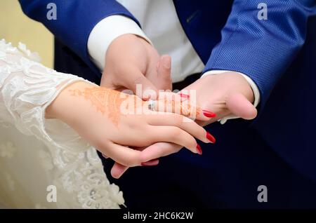 Ein Bräutigam legt einen Ehering auf die Hand einer arabischen Braut, die ihr Henna auf die Hände legt Stockfoto