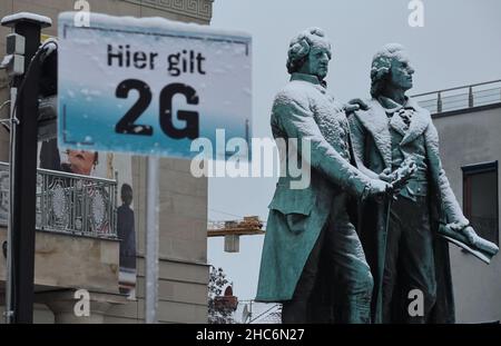 Weimar, Deutschland. 25th Dez 2021. Das Goethe-Schiller-Denkmal am Theaterplatz ist mit Schnee bedeckt. Davor weist ein Schild auf der Eisbahn auf die Corona-Regel von 2G hin. Die bronzene Doppelstatue vor dem Deutschen Nationaltheater wurde im 19th. Jahrhundert vom Dresdner Bildhauer E. Rietschel entworfen. Quelle: Soeren Stache/dpa-Zentralbild/dpa/Alamy Live News Stockfoto