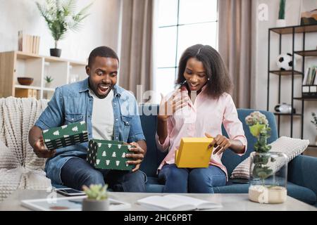 afroamerikanischer Mann und Frau mit echtem Glück auf ihrem Gesicht öffnen bunte Geschenk-Boxen. Angenehmes junges Paar, das Geschenke teilt, während es auf der bequemen Couch sitzt. Stockfoto