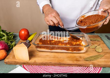 Eine unkenntliche Frau, die ein Lasagne-Rezept macht, indem sie Bolognese-Sauce mit den verschiedenen Zutaten um den Contai auf die Zubereitung setzt Stockfoto