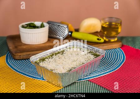 Ungekochte Lasagne mit Pesto und geriebenem Käse in einem Folientablett mit einigen Zutaten und Geschirr dahinter auf einem Tisch. Stockfoto