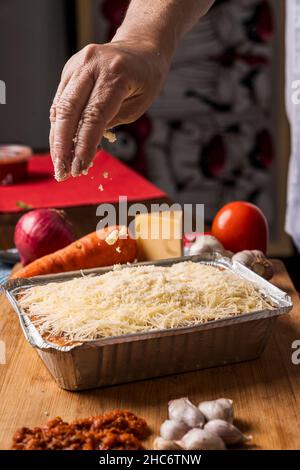 Vertikales Bild einer Hand einer nicht erkennbaren Person, die viel geriebenen Käse über Lasagne mit verschiedenen Arten von Zutaten um sie herum gießt. Stockfoto