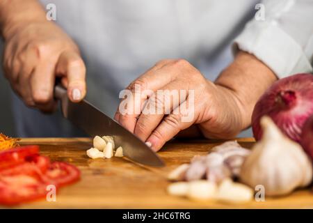 Nahaufnahme der Hände einer erwachsenen Frau, die mit einem Küchenmesser einen Knoblauch auf ein Schneidebrett hackt, um Lasagne vorzubereiten. Stockfoto