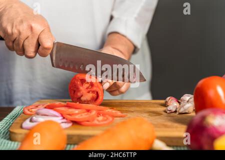 Nahaufnahme der Hände einer erwachsenen Frau, die Tomaten mit einem Küchenmesser auf einem hölzernen Schneidebrett zur Lasagne-Zubereitung in Scheiben schneidet. Stockfoto