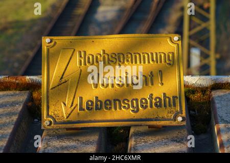 Schild: Hochspannung, Todesgefahr (deutsch: Hochspannung, Vosicht Lebensgefahr), mit Eisenbahnschienen im verschwommenen Hintergrund Stockfoto