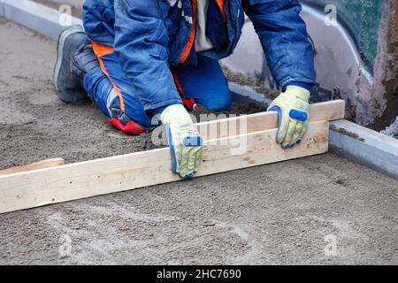 Ein kniender blauer Overalls-Arbeiter ebnet ein sandiger Boden mit einer Holzebene für zukünftige Gehwegverkleidungen. Speicherplatz kopieren. Stockfoto