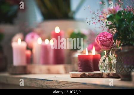 Selektive Fokusaufnahme einer rosa Rose in einem kleinen Glas neben Pflanzen und zündete Kerzen auf einer Party an Stockfoto