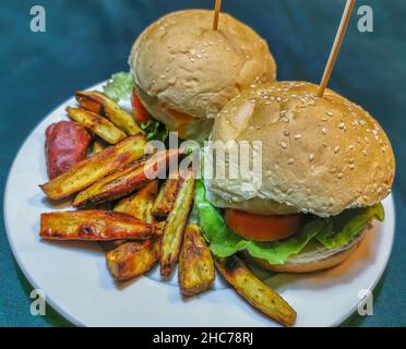Blick auf zwei Veggie-Burger auf dem Teller, auf einem Brötchen mit Sesam, serviert mit traditioneller gebratener Süßkartoffel... Stockfoto