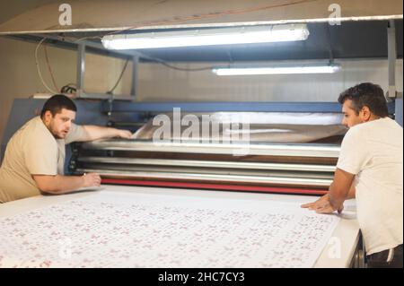 Zwei Techniker Arbeiter Betreiber arbeitet an großen Premium-Gewebe industriellen Textil Sublimationswalze Heat Press Druckmaschine in der digitalen Druckerei o Stockfoto