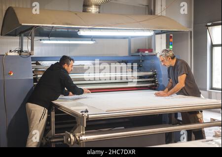 Zwei Techniker Arbeiter Betreiber arbeitet an großen Premium-Gewebe industriellen Textil Sublimationswalze Heat Press Druckmaschine in der digitalen Druckerei o Stockfoto