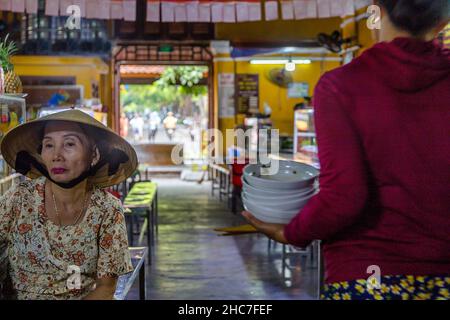 Eine Dame mit einem konischen Hut sitzt an der Theke, während ein Servicemitarbeiter Geschirr trägt. Stockfoto