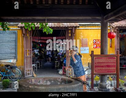 Eine vietnamesische Dame zieht den Eimer aus dem Brunnenwasser vor dem Eingang. Das Hotel liegt vor dem Central Market, der Altstadt, Hoi an. Stockfoto