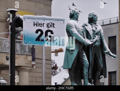 Weimar, Deutschland. 23rd Dez 2021. Das Goethe-Schiller-Denkmal am Theaterplatz ist mit Schnee bedeckt. Auf der linken Seite weist ein Schild an der Eisbahn auf die Ordonanzregel von 2G hin. Die bronzene Doppelstatue vor dem Deutschen Nationaltheater wurde im 19th. Jahrhundert vom Dresdner Bildhauer Ernst Rietschel entworfen. Quelle: Soeren Stache/dpa-Zentralbild/ZB/dpa/Alamy Live News Stockfoto