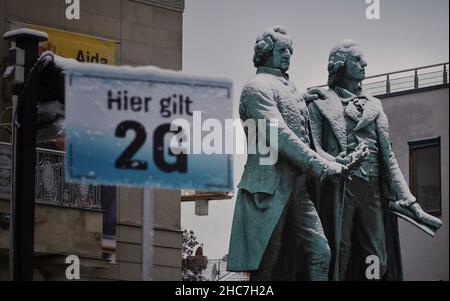 Weimar, Deutschland. 23rd Dez 2021. Das Goethe-Schiller-Denkmal am Theaterplatz ist mit Schnee bedeckt. Auf der linken Seite weist ein Schild an der Eisbahn auf die Ordonanzregel von 2G hin. Die bronzene Doppelstatue vor dem Deutschen Nationaltheater wurde im 19th. Jahrhundert vom Dresdner Bildhauer Ernst Rietschel entworfen. Quelle: Soeren Stache/dpa-Zentralbild/ZB/dpa/Alamy Live News Stockfoto