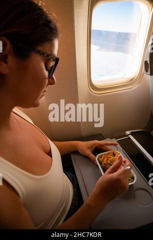 Kaukasische Frau, die in der Economy Class fliegt und an Bord des Flugzeugs ein Mittagessen von einem Tablett isst. Stockfoto