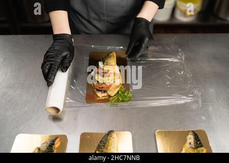 Frau verwendet Frischhaltefolie, um Essen auf dem Tisch in einem Restaurant in der Küche zu lagern. Eine Rolle transparenter Kunststoff-Frischhaltefolie für Lebensmittelverpackungen. Gebacken Stockfoto