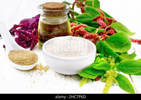 Amaranth-Mehl in einer Schüssel, die Samen im Löffel und das Öl im Glas, braune, grüne und violette Blüten einer Pflanze auf dem Hintergrund des Holzbretts Stockfoto