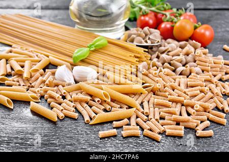 Pasta Vollkorn und Roggenpfeife rigate, Spaghetti, Penne; Muscheln, Rigatoni, Tomaten, Knoblauch, Pflanzenöl in einem Dekanter auf dunklem Holzbrett Stockfoto