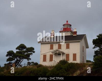 Zweistöckiger Leuchtturm aus weißem Holz in der Yaquina Bay mit verschalzten Fenstern und rotem Metallrahmen über dem Leuchtfeuer des Leuchtturms. Stockfoto