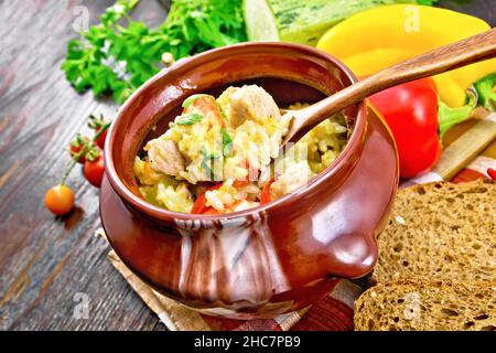 Reis mit Huhn, Paprika, Zucchini und Karotten in einem Tontopf mit einem Löffel auf Serviette auf Holzbrett Hintergrund Stockfoto