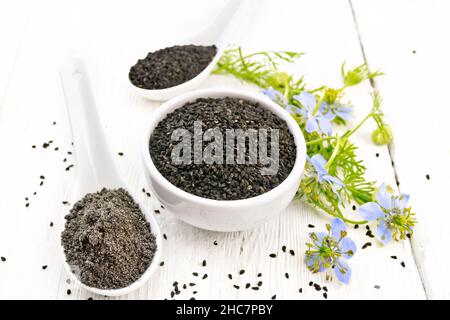 Schwarze Kümmel-Samen in einer Schüssel, Mehl und Samen in Löffeln, Kalingi-Zweige mit blauen Blumen und Blättern auf einem Holzbrett Hintergrund Stockfoto