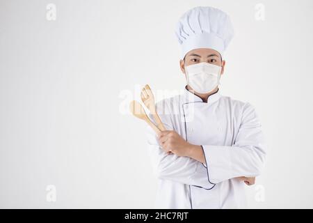 Portrait des Restaurantkochs in weißer Uniform und medizinischer Maske mit hölzernen Kochspateln Stockfoto