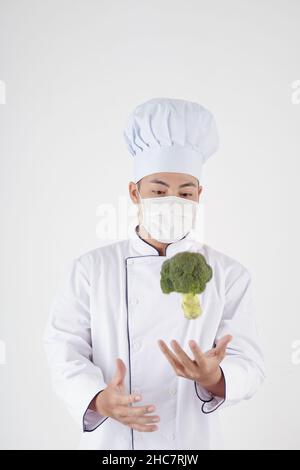 Restaurantchef in medizinischer Maske wirft Brokkoli, isoliert auf weiß Stockfoto