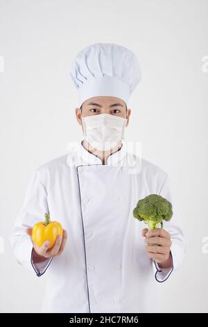 Portrait des Cafe-Küchenchefs in Hut und Schutzmaske mit Paprika und Brokkoli Stockfoto