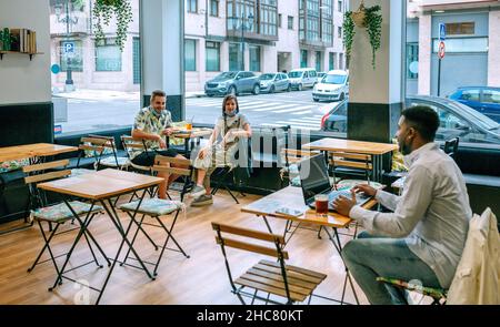 Junger Mann, der in einem Café Telearbeit mit Freunden unterhält, die soziale Distanz bewahren Stockfoto